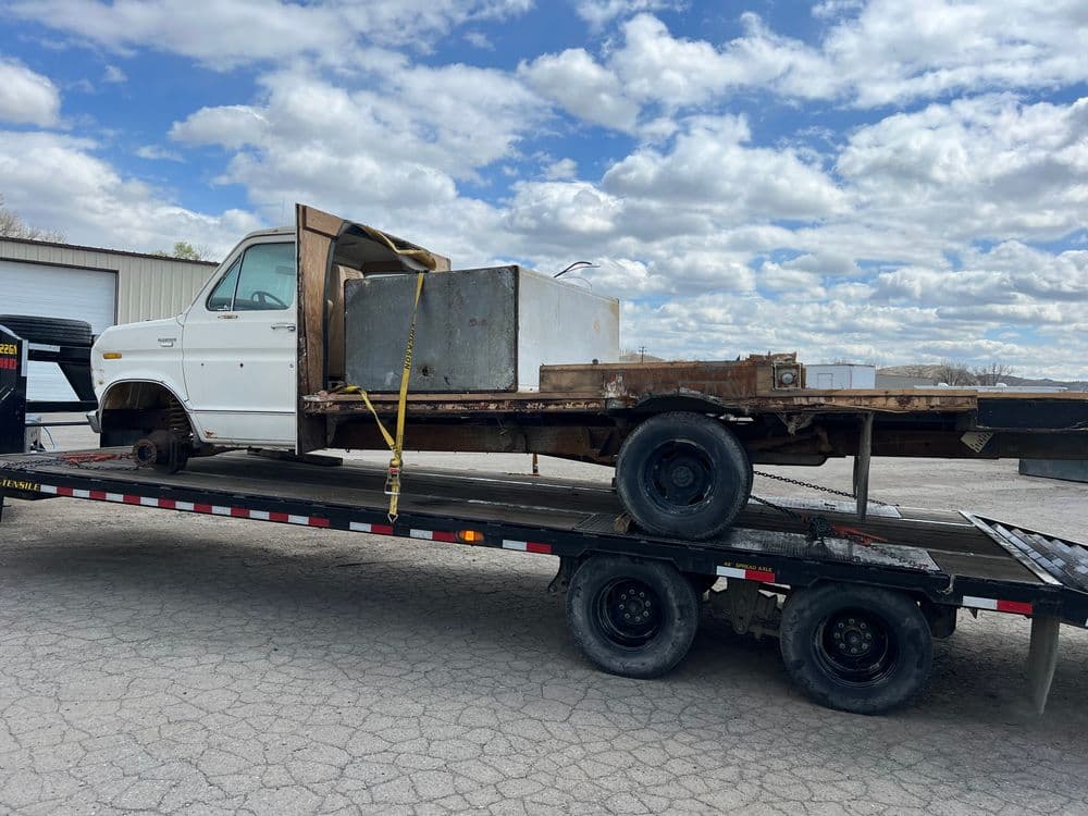 Old truck on trailer with metal box, cloudy sky in background, construction site setting.