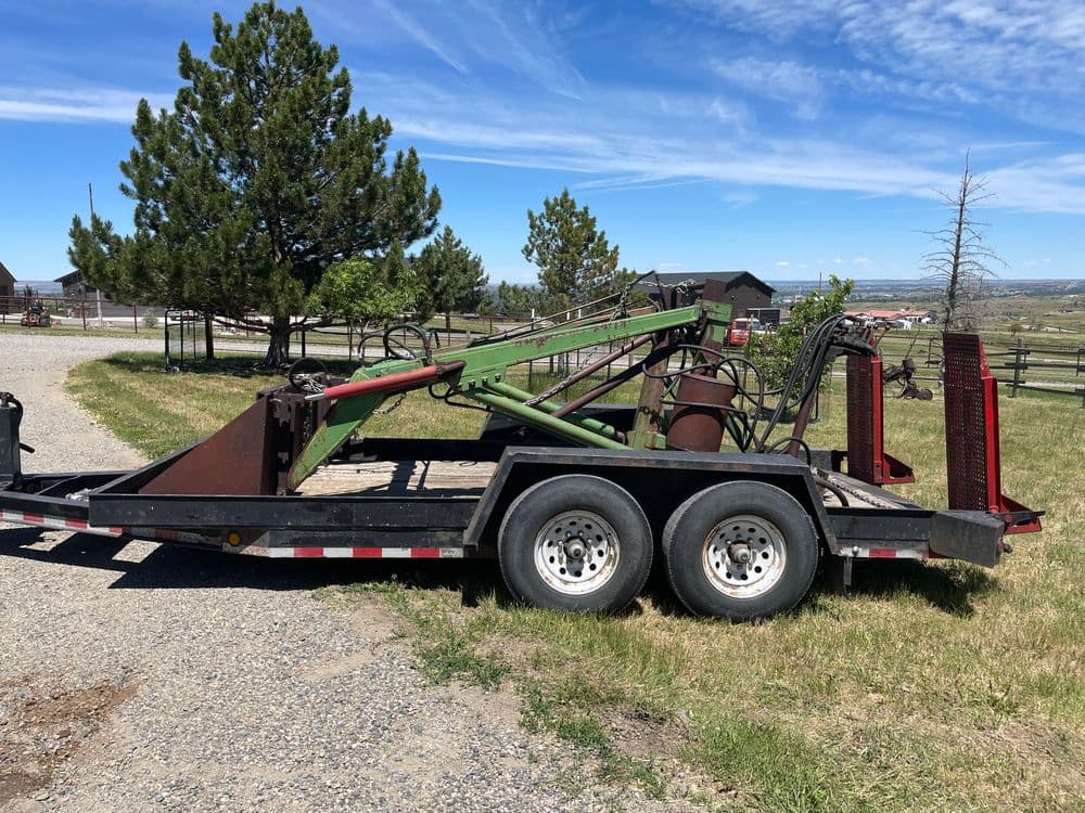 Green farming equipment on a trailer in a rural landscape with trees and buildings.