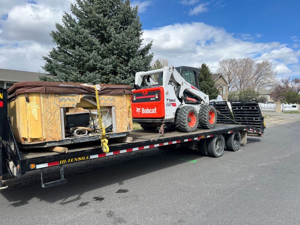 Bobcat skid steer on flatbed trailer alongside a discarded hot tub and debris.
