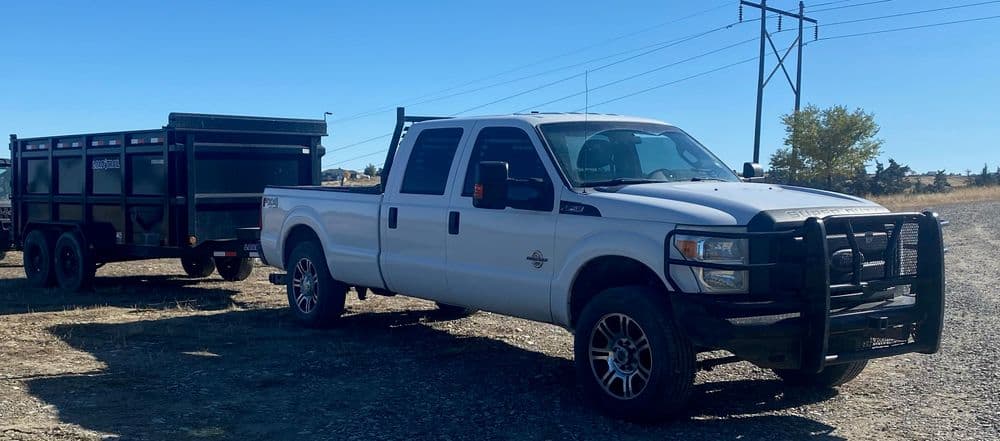 White Ford F-250 truck with a trailer parked on a gravel road under a clear blue sky.