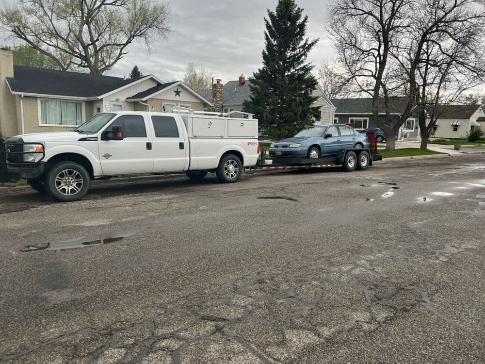 White pickup truck towing a blue car on a trailer in a residential neighborhood.