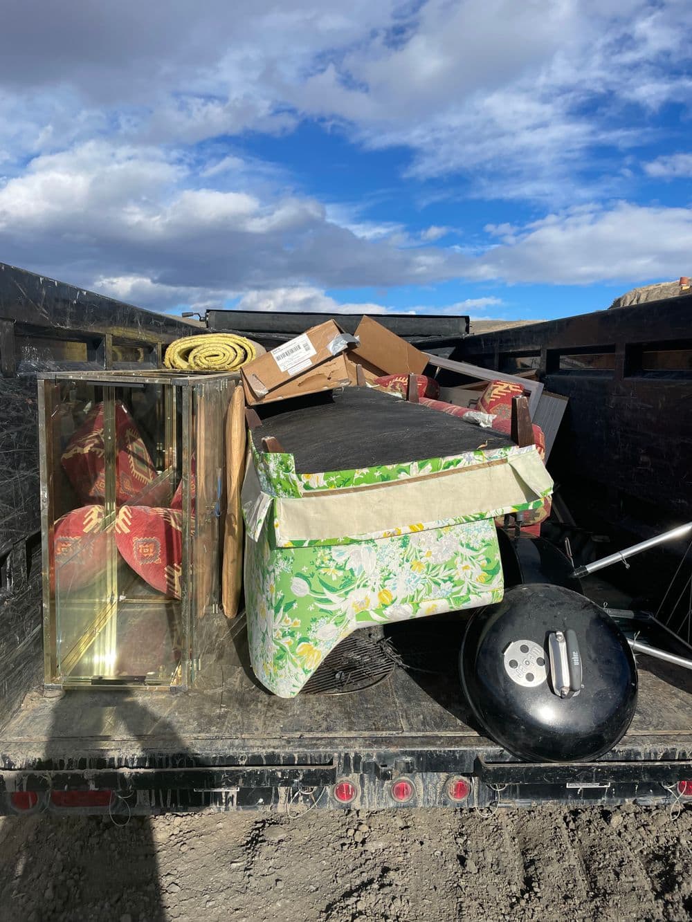 Cluttered truck bed filled with furniture, boxes, and a barbecue grill against a cloudy sky.