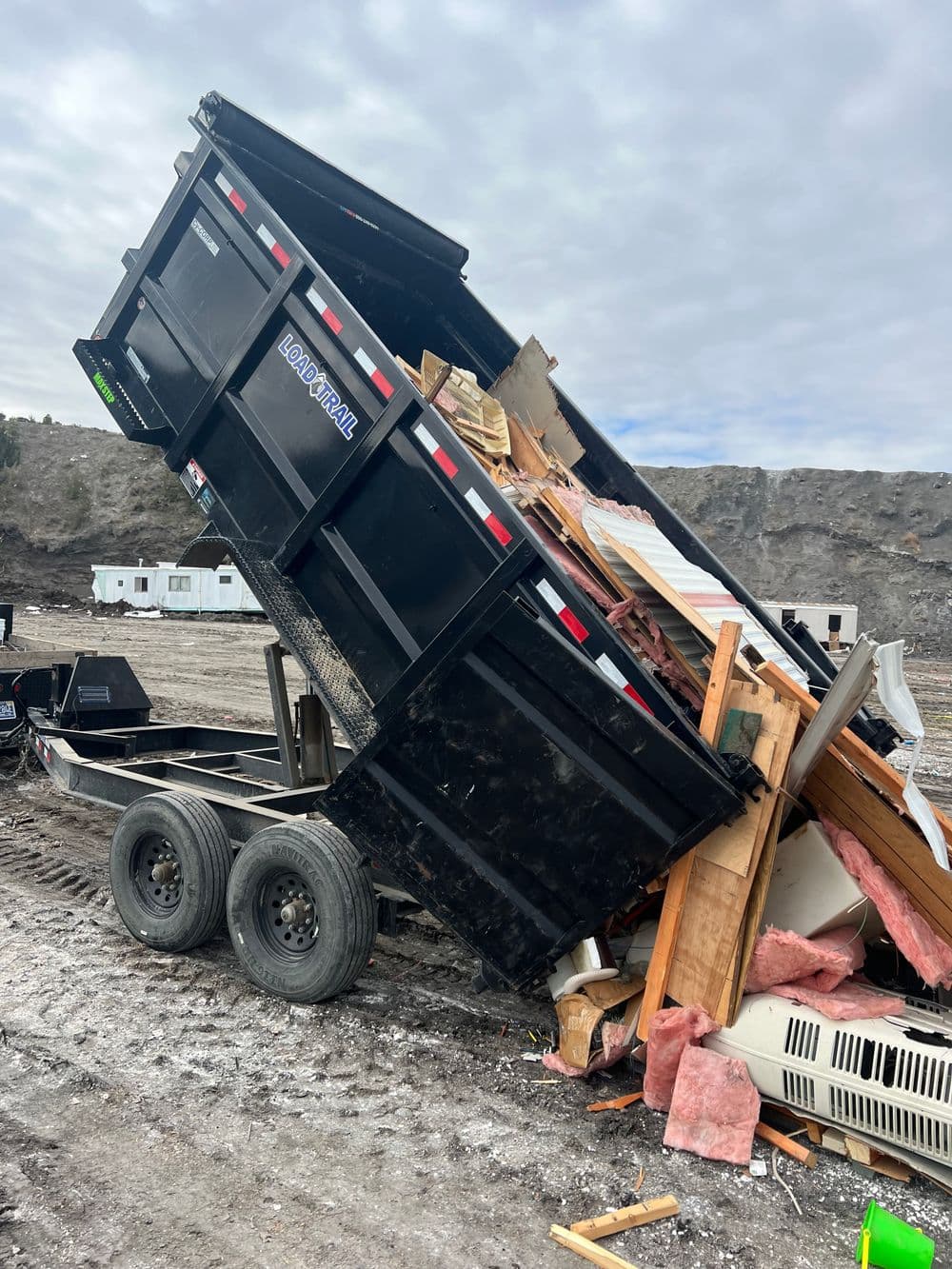 Black dump trailer unloading construction debris at a job site with cloudy sky.