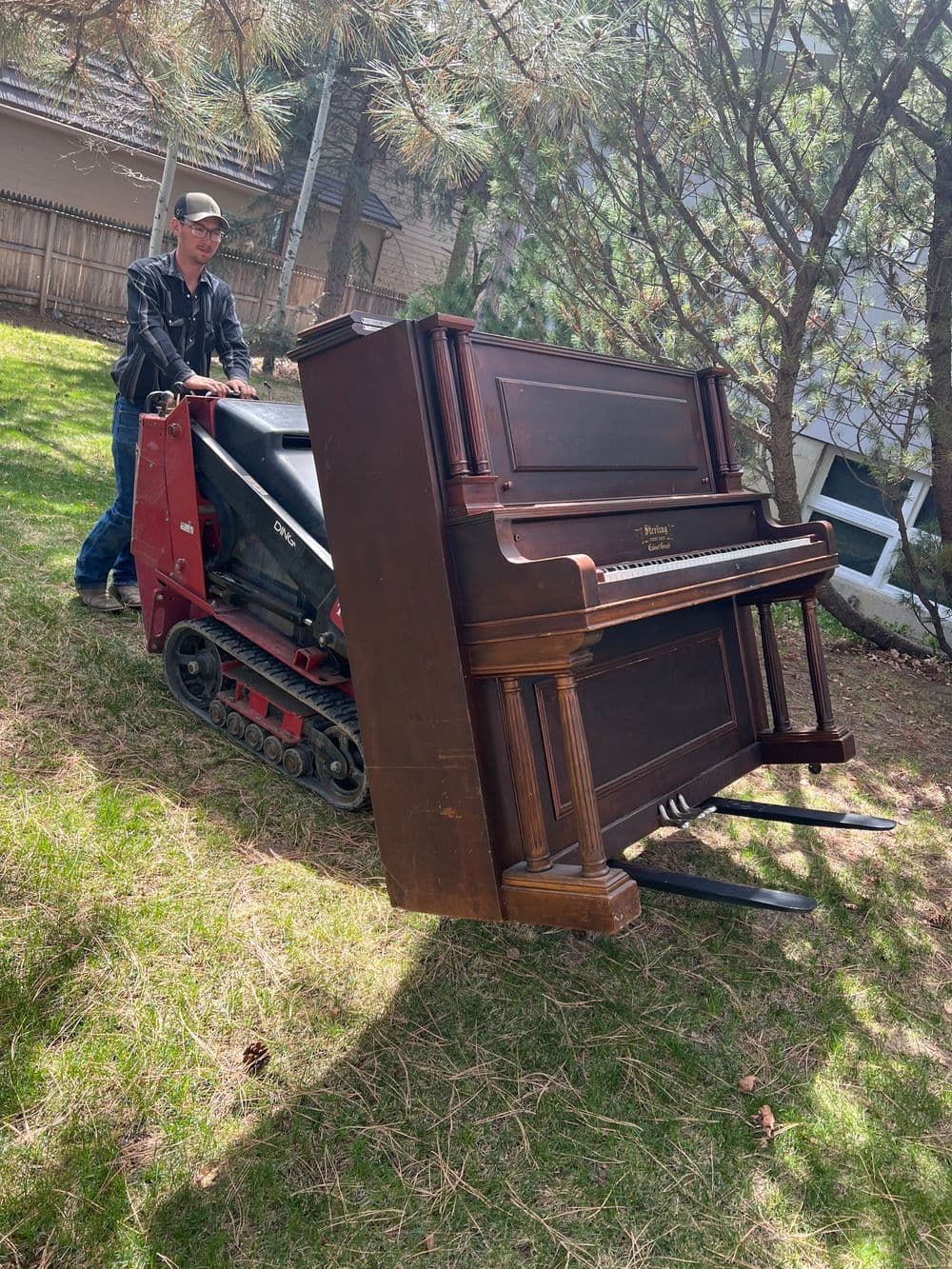 Man using a tracked vehicle to move a wooden piano across a grassy area.