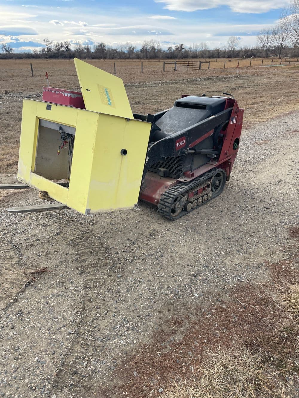 Red tracked machine with a yellow box attachment on gravel farm road in open field.