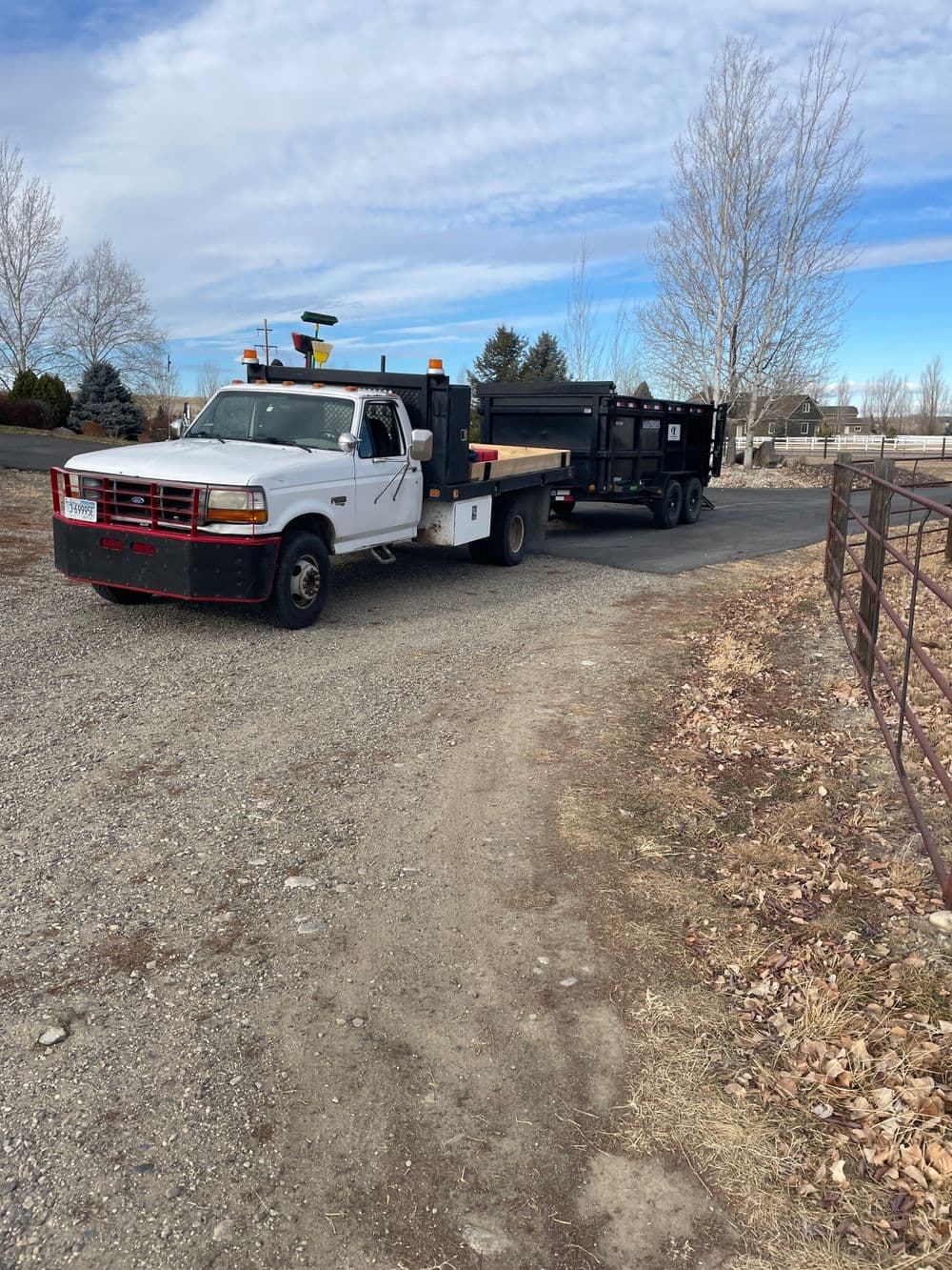 White truck towing a black trailer on a gravel road with trees and blue sky in the background.