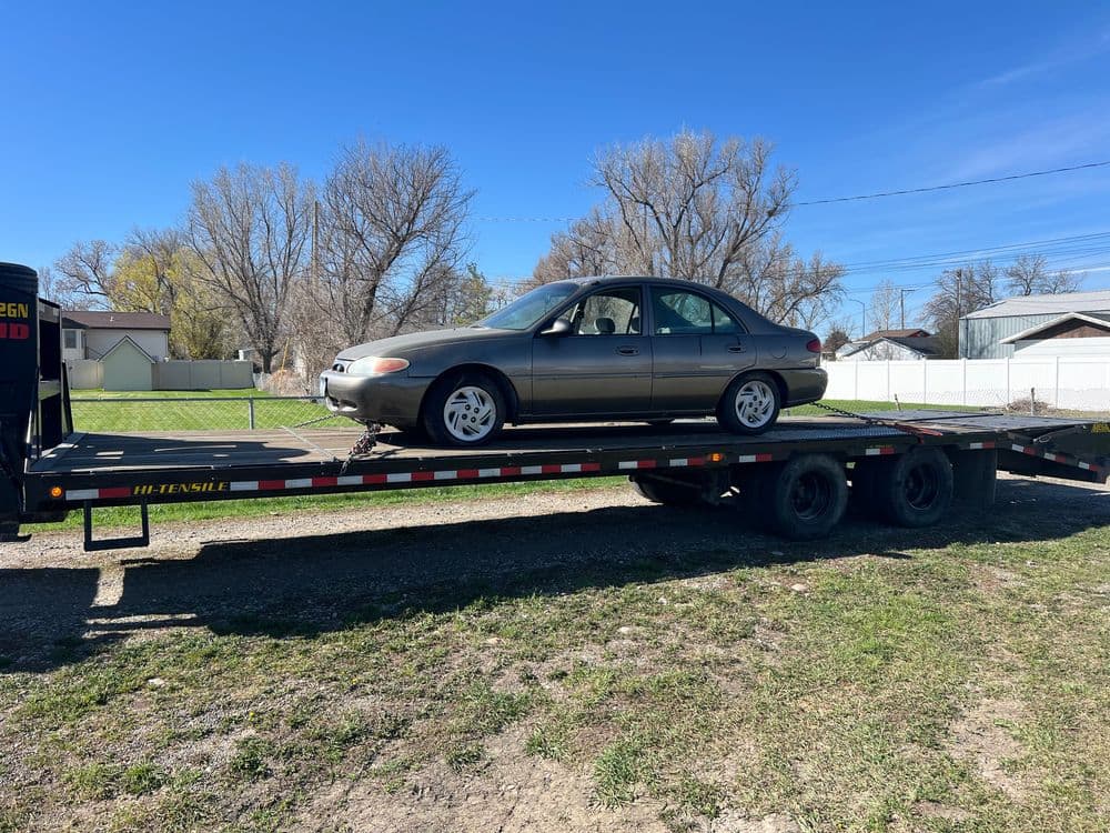 Brown sedan being transported on a flatbed tow truck against a clear blue sky.